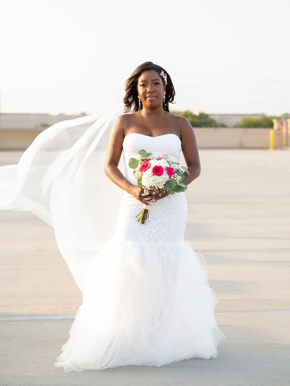 A bride in a flowing white strapless wedding gown holds a pink and white bouquet outdoors at sunset.