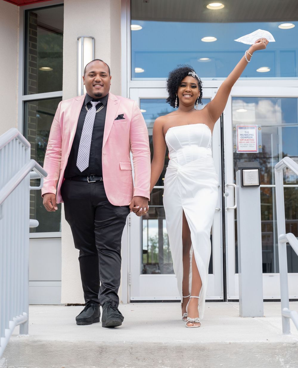 A joyful couple celebrates their wedding day outside, with the groom in a pink blazer and bride in white dress holding paper up.