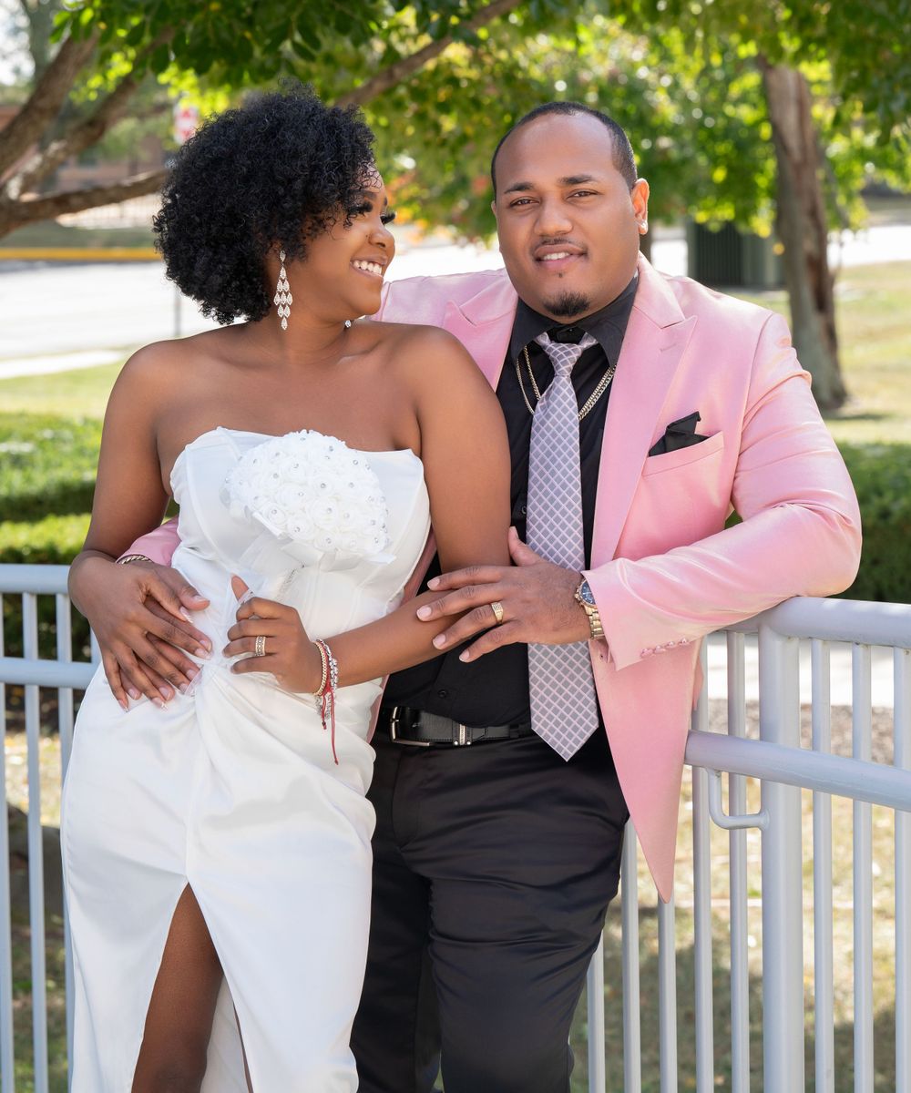 Wedding couple in pink suit and white dress posing outdoors on sunny day.