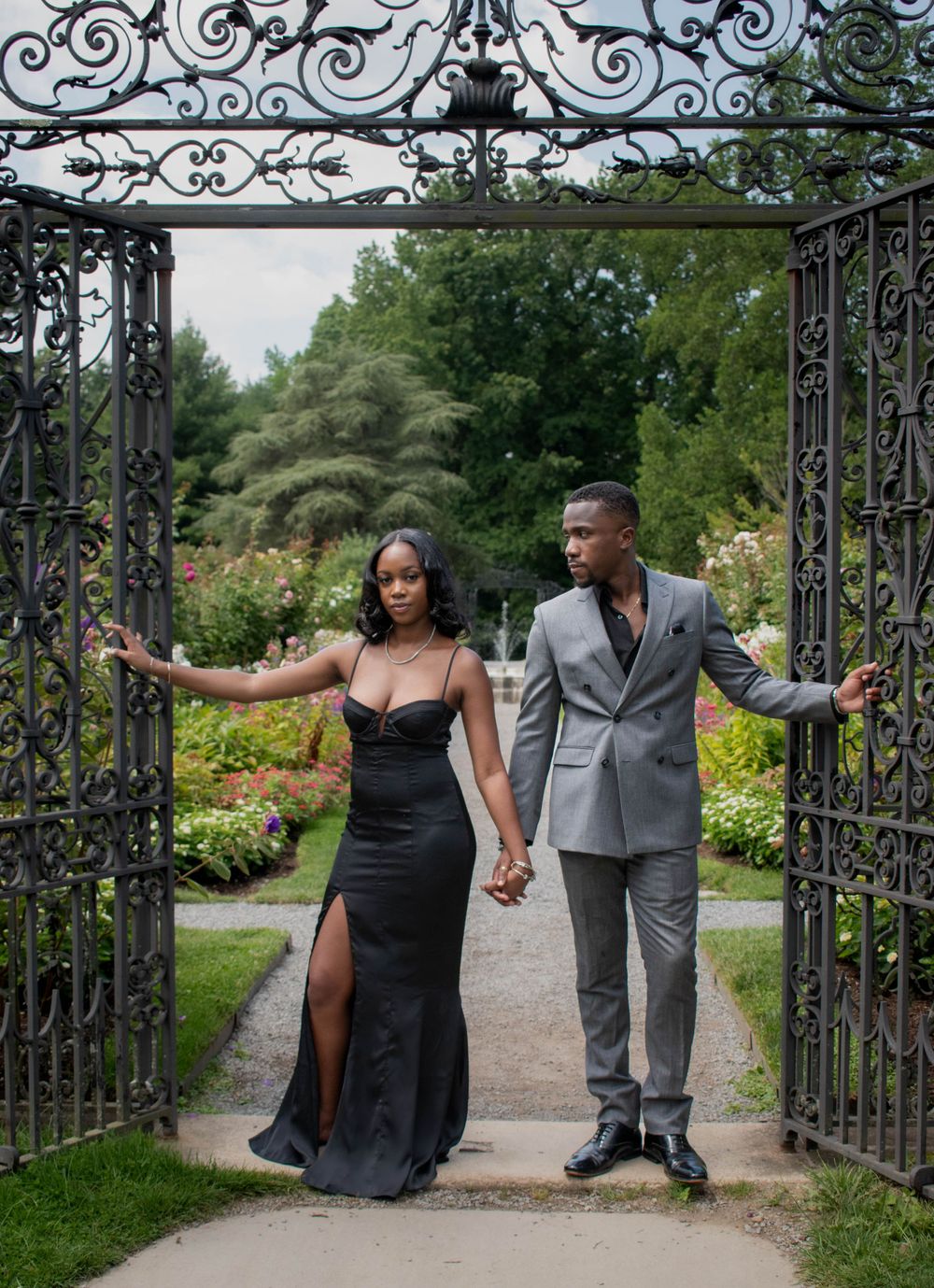 A romantic couple pose in formal attire beneath an ornate iron garden gate archway.