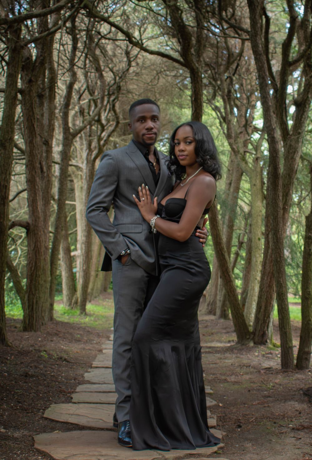 Romantic couple in formal attire embrace on a wooded path surrounded by tall trees.