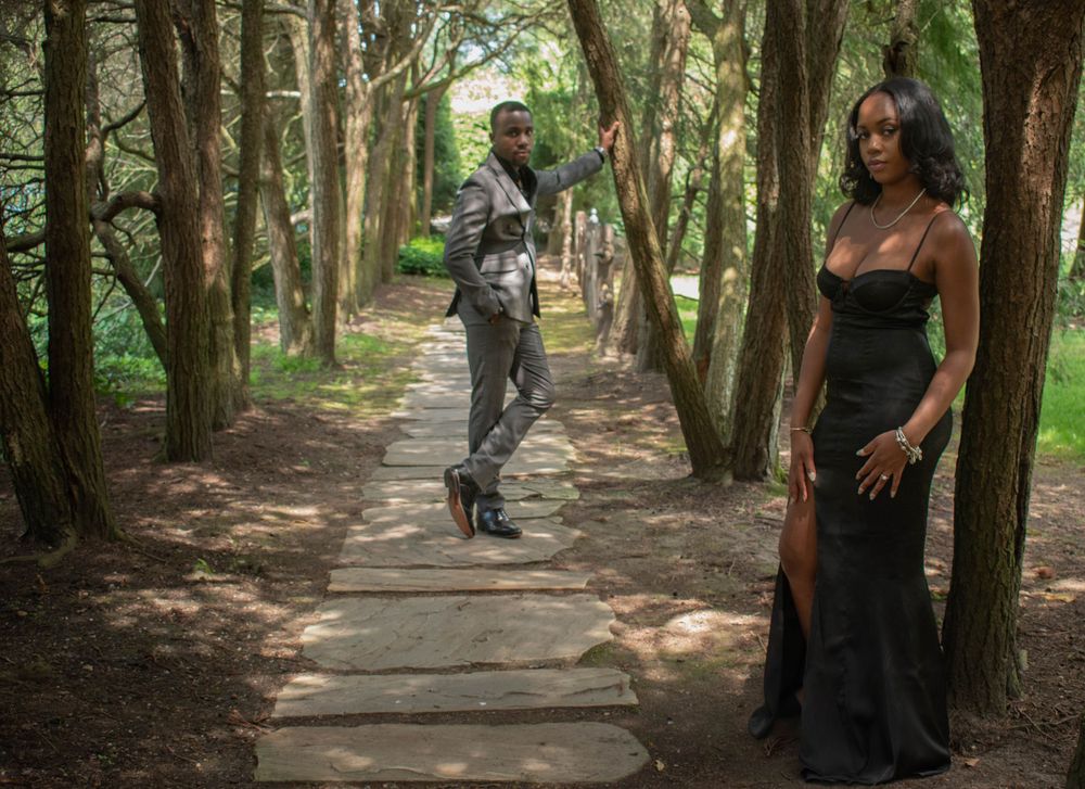A stylish couple in formal wear walking down a tree-lined garden path.