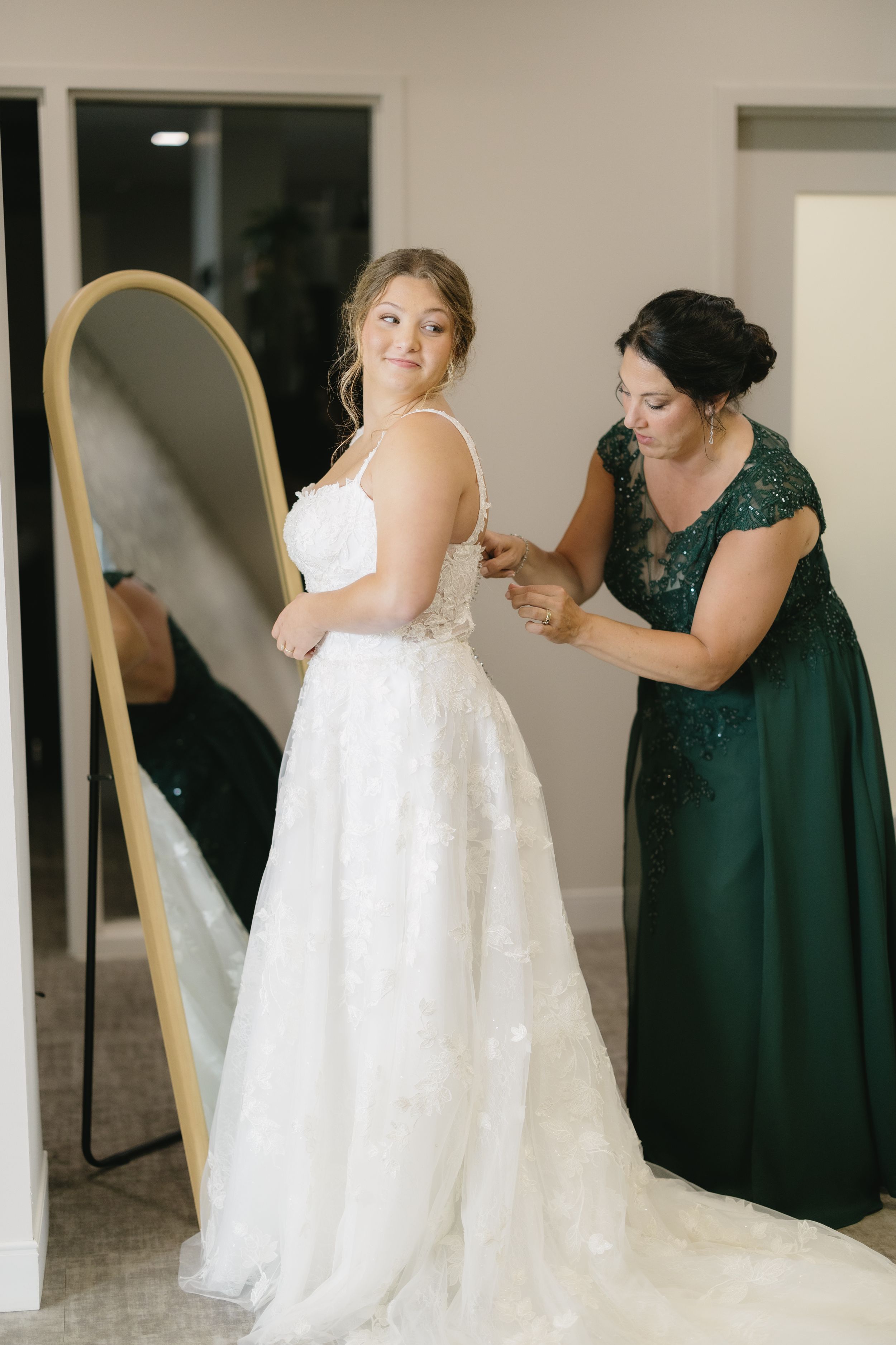 Bridesmaids in emerald green dresses help bride with white lace wedding gown during wedding preparations.