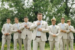 A group of groomsmen in light beige suits pose together outdoors with trees in the background.