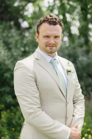 A groom wearing a light beige suit and mint green tie poses outdoors in a garden setting with greenery in the background.
