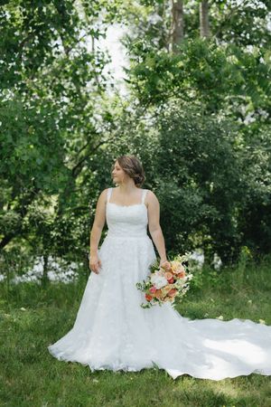 A bride in a flowing white lace gown holds a peach and coral bouquet while standing in a sunlit garden setting.