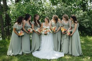 A bridal party wearing sage green bridesmaid dresses holds peach and orange bouquets while posing outdoors.