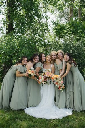 A wedding party in sage green bridesmaid dresses poses together with vibrant peach floral bouquets in a garden setting.