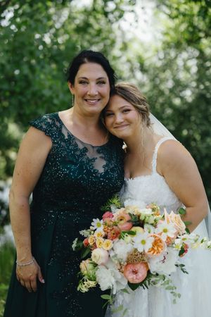 Two women in forest greenery, one wearing an emerald lace gown and one in white wedding dress holding peach floral bouquet.