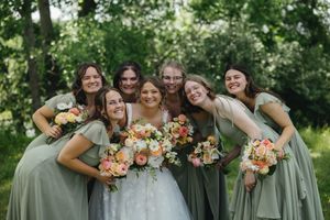 A group of bridesmaids in sage green dresses pose with colorful peach and orange bouquets outdoors.