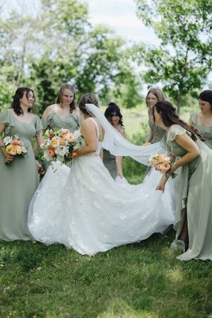 Bridesmaids in sage green dresses help adjust bride's white lace wedding gown while holding peach bouquets outdoors.