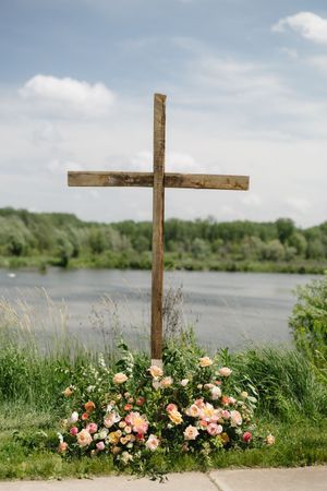 A wooden cross stands on a grassy riverbank with pink roses arranged at its base against a serene water landscape.