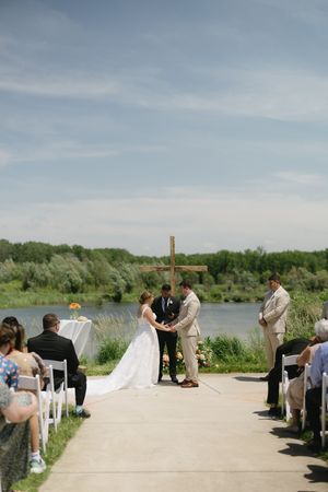 A wedding ceremony takes place on a paved path beside a serene lake with greenery and a wooden cross in the background.