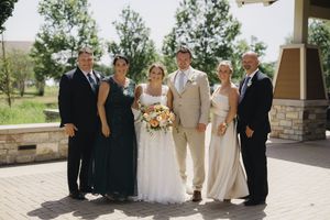 A group in formal attire poses outdoors at a wedding ceremony with stone pillars and greenery in the background.