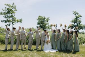 Wedding party in sage green attire stands together for outdoor group photos in a sunny field.