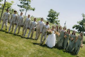 Wedding party in light gray and white attire poses together on a grassy hillside during outdoor summer celebration.