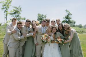 Wedding party wearing sage green and beige attire poses together outdoors in a joyful group photo.