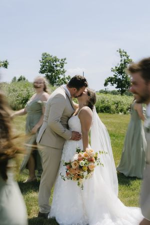 Two people sharing a romantic kiss at an outdoor wedding ceremony with bridesmaids in sage green dresses and peach floral bouquet.