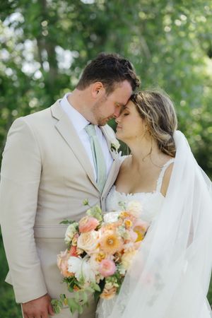 A romantic wedding couple embrace outdoors with the bride holding a soft peach and pink floral bouquet.