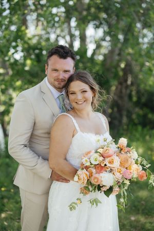 A joyful couple poses outdoors in wedding attire with the bride holding a peach and cream colored floral bouquet.