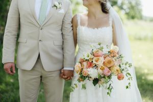Wedding details showcased with a beige suit and white dress adjacent to a peach and coral floral bouquet in an outdoor setting.