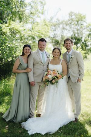 Wedding party poses outdoors in sage green dresses and tan suits on a sunny summer day surrounded by greenery.