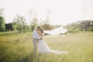 A romantic wedding couple embraces in a sunlit meadow as the bride's veil flows gracefully in the breeze.