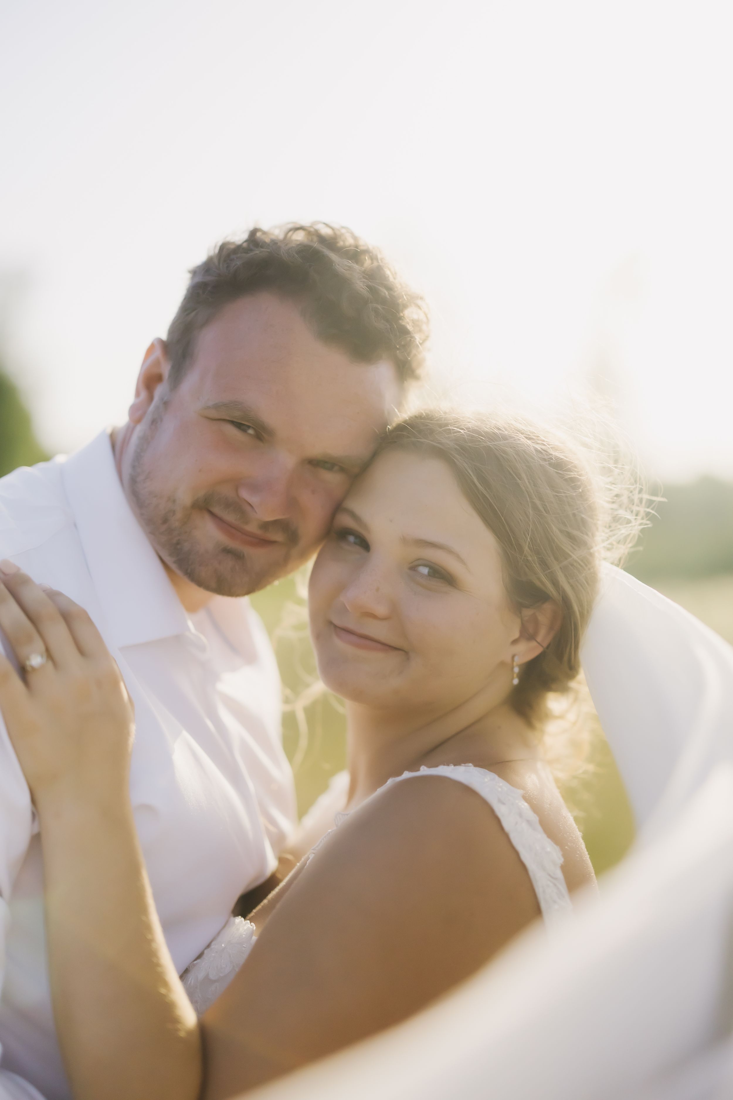 Romantic newlywed couple embraces outdoors during golden hour with sunlight creating a dreamy backlit effect.