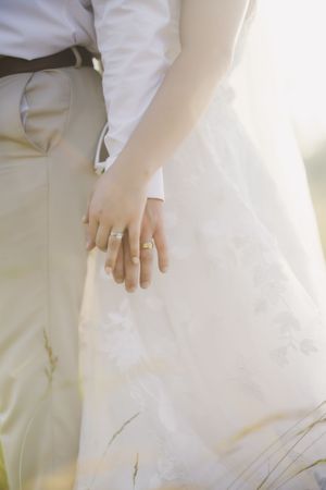 Close-up shot of a couple's hands with wedding rings, with the bride wearing a white dress and groom in light colored attire.