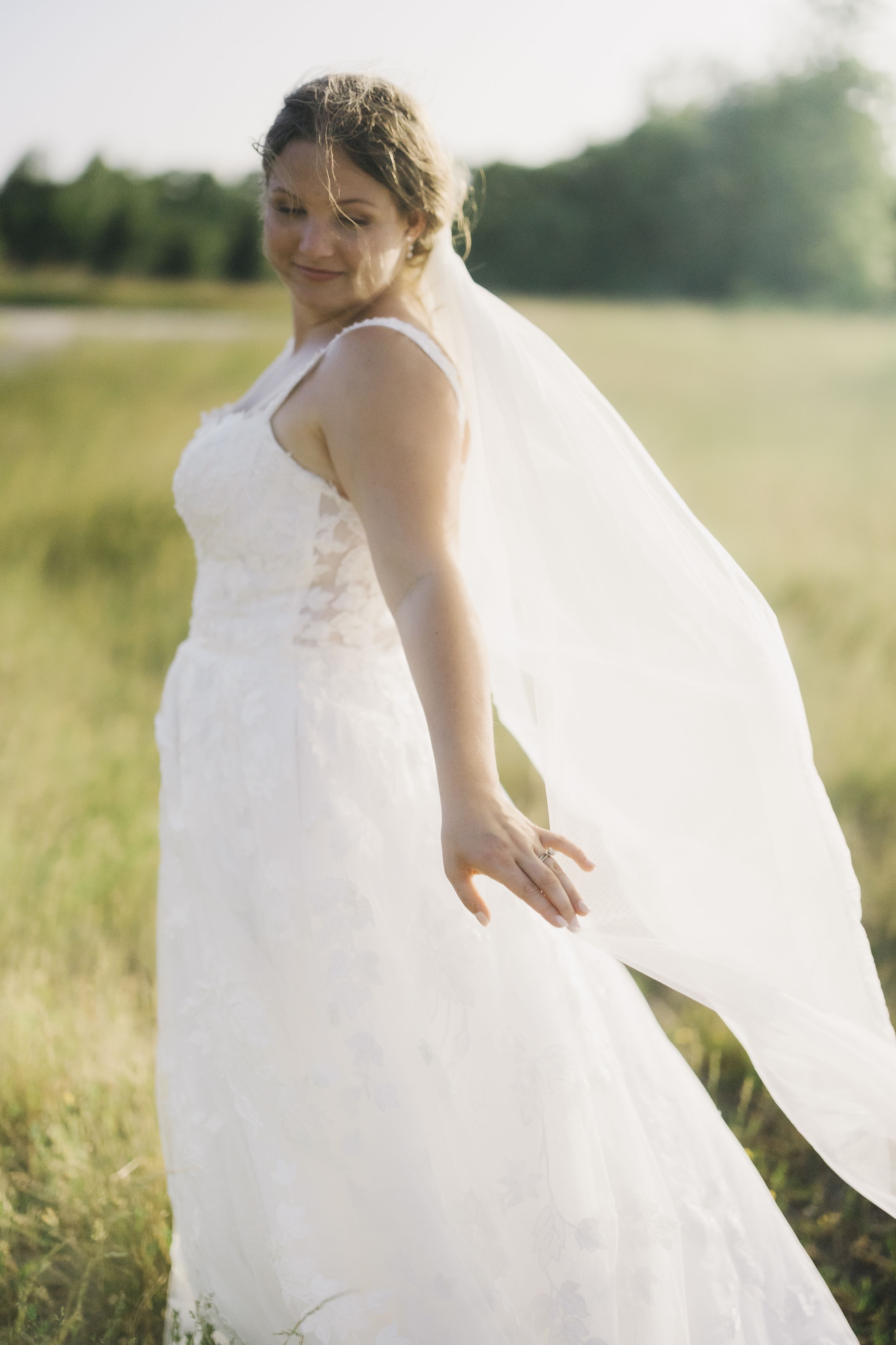 A wedding gown with flowing veil catches sunlight in a grassy meadow during golden hour.