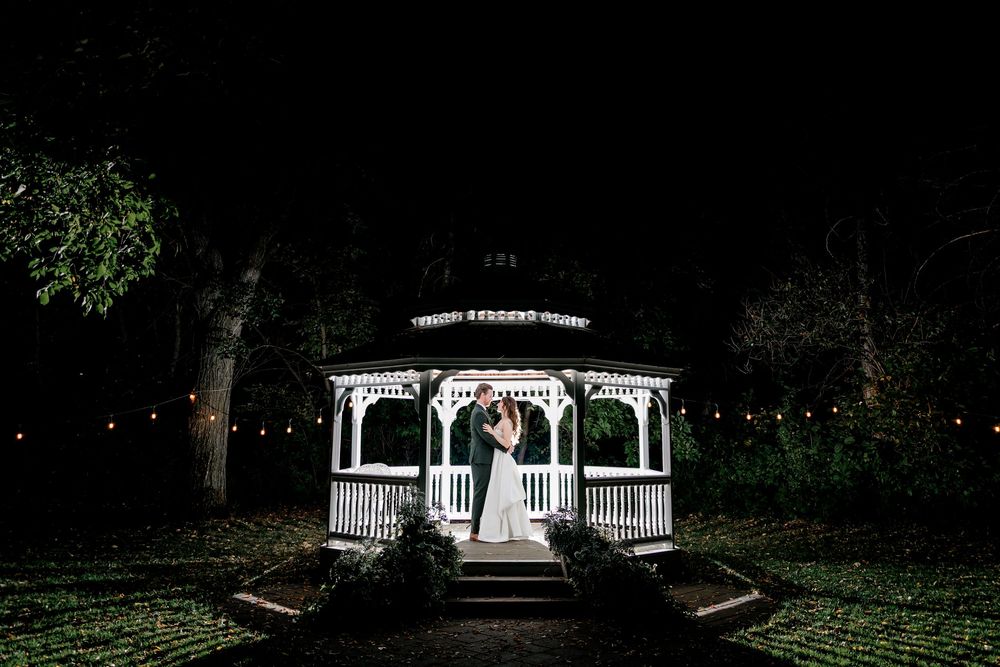 A white gazebo illuminated at night with trees and landscaping in the background.