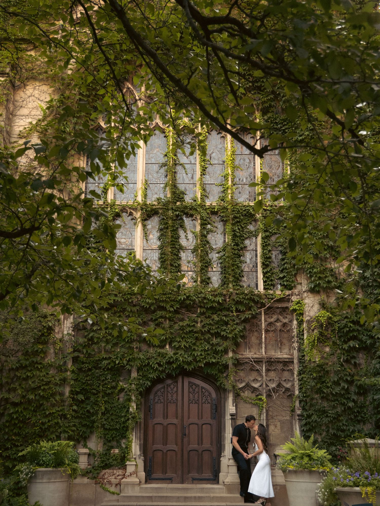 Couple embracing by an ivy-covered Gothic building entrance with wooden doors.
