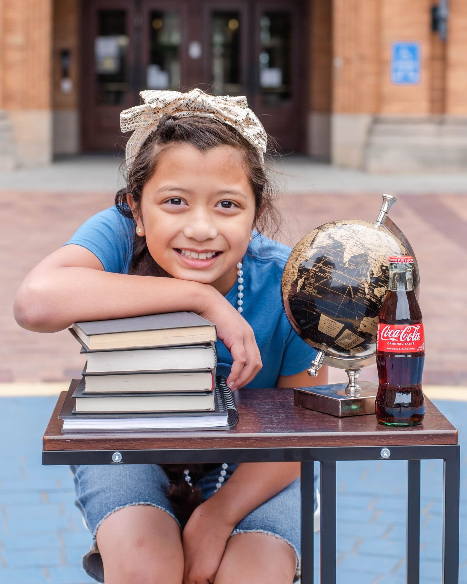 Child poses with stack of books and Coca-Cola bottle on outdoor table.