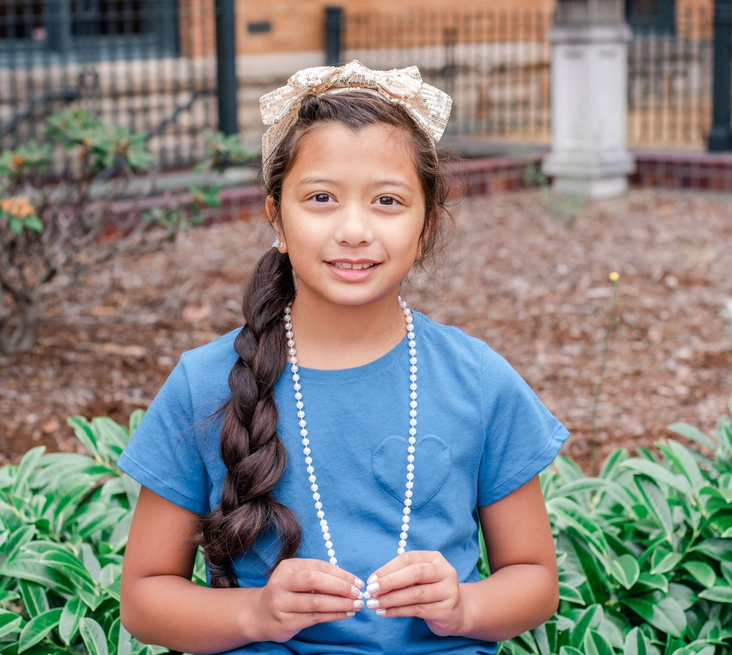 Young student in blue shirt wearing pearl necklace smiles while sitting outdoors near brick building.
