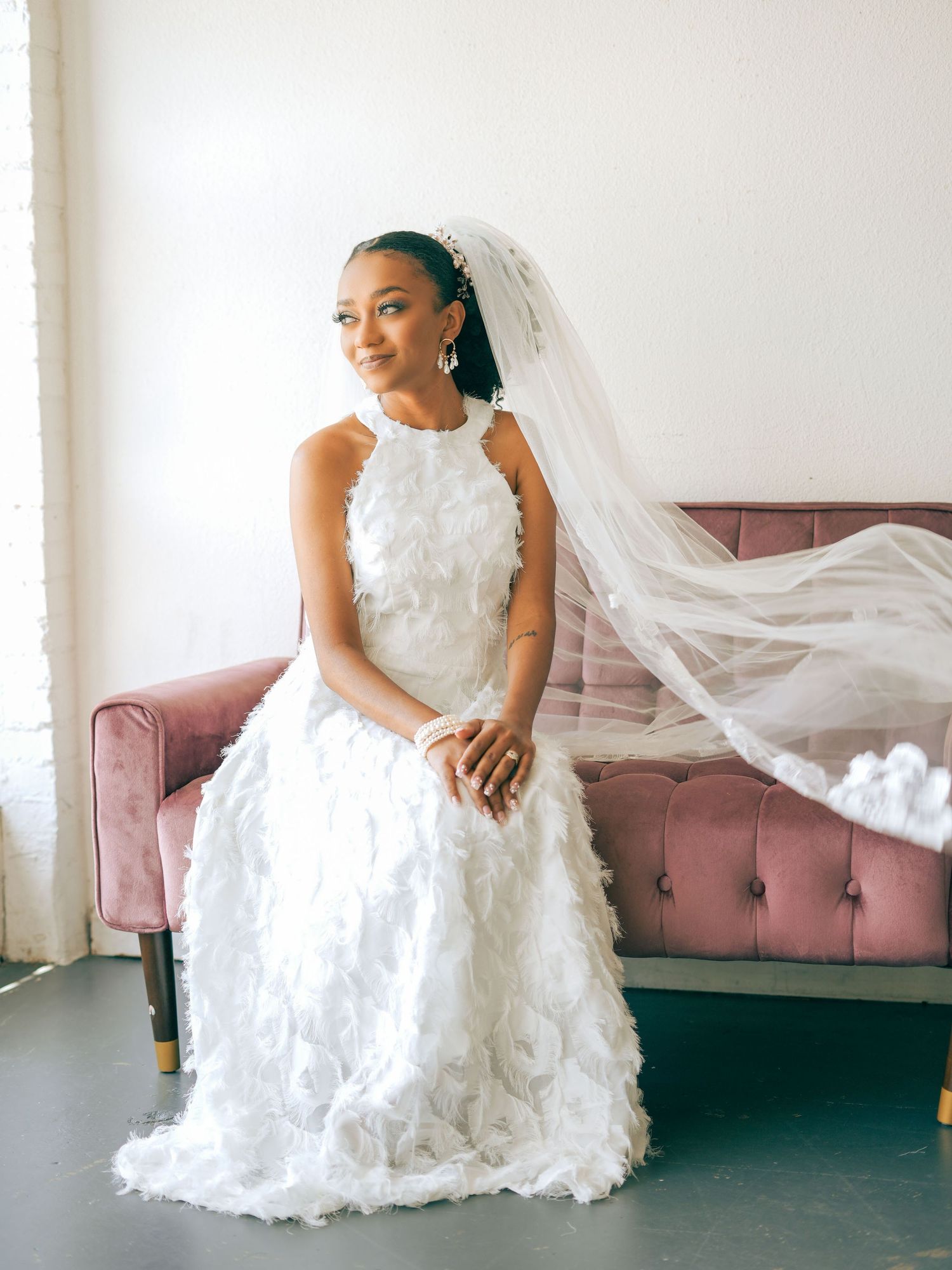 Bride in textured white gown with flowing veil sits elegantly on pink tufted sofa.