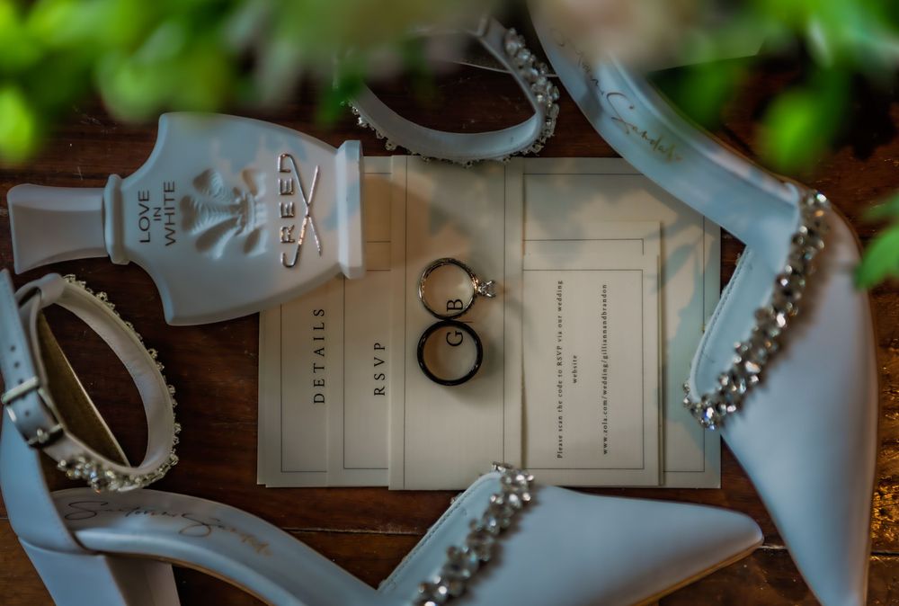 Wedding flat lay with Creed perfume, diamond rings, heels, pearl bracelet, and RSVP cards on wood.