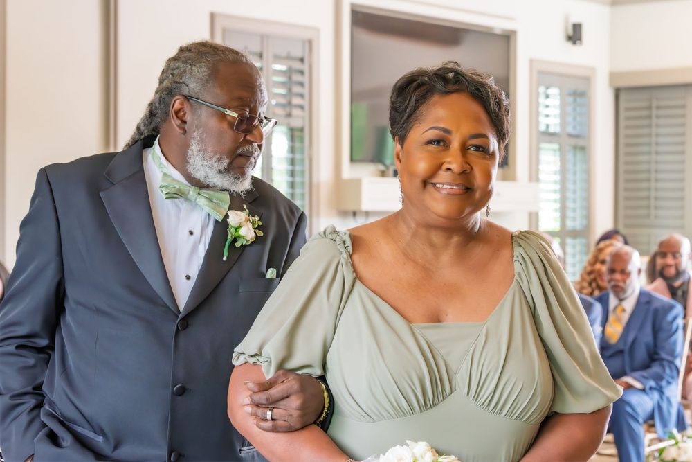 Elegant couple in sage green attire walks arm-in-arm at a formal wedding ceremony indoors.