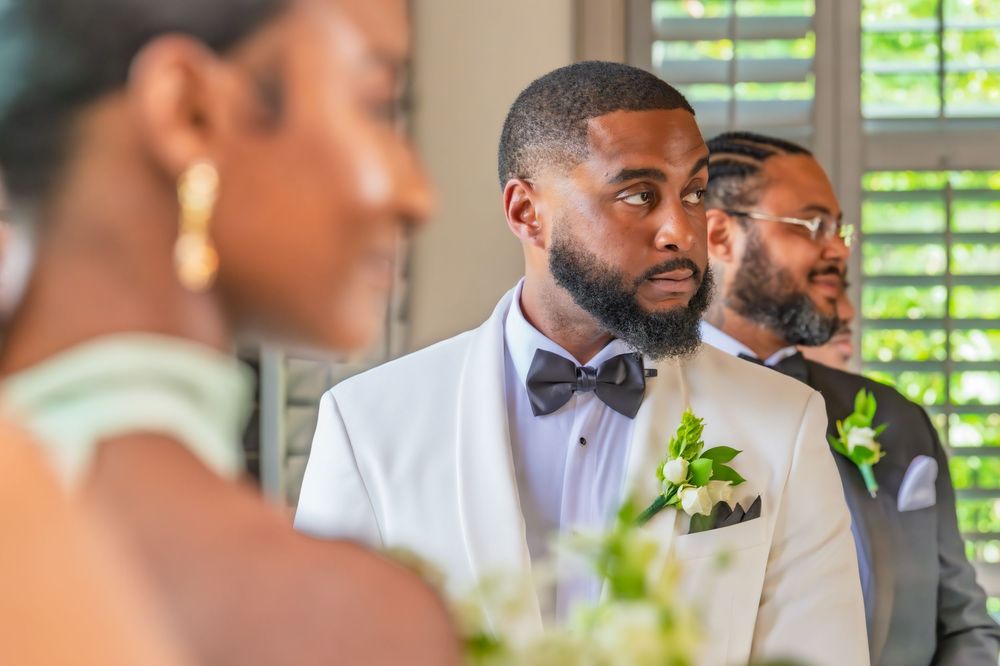 Groom in white tuxedo with black bow tie looks emotional during wedding ceremony with groomsmen behind him.
