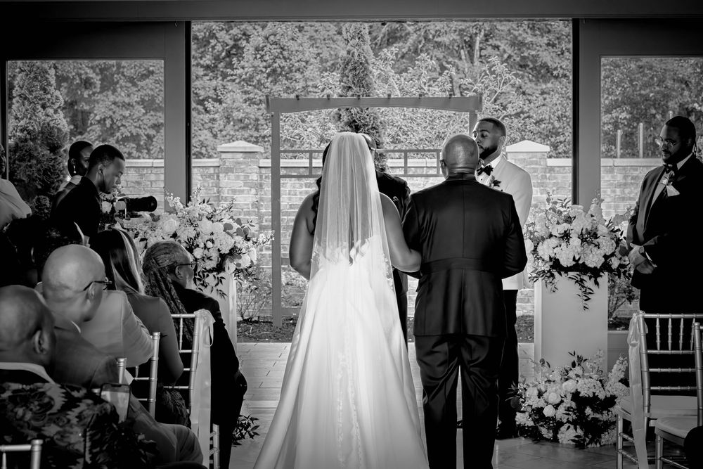 Black and white wedding ceremony photo showing bride in white gown and veil walking down aisle with escort.