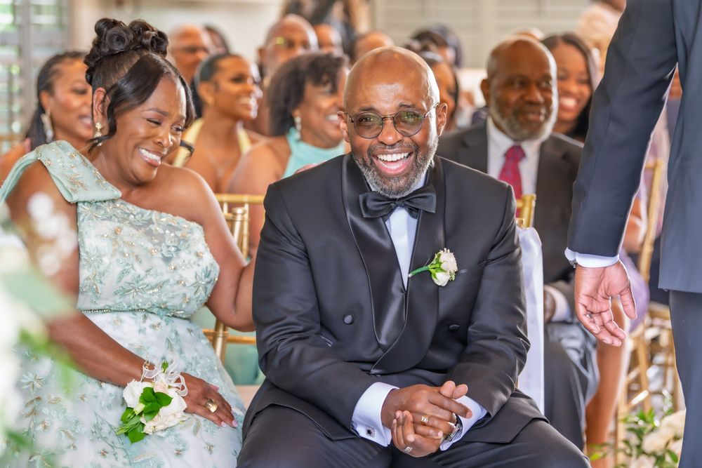 Joyful wedding guests, a smiling couple in formal attire, seated at an elegant ceremony with guests laughing in background.