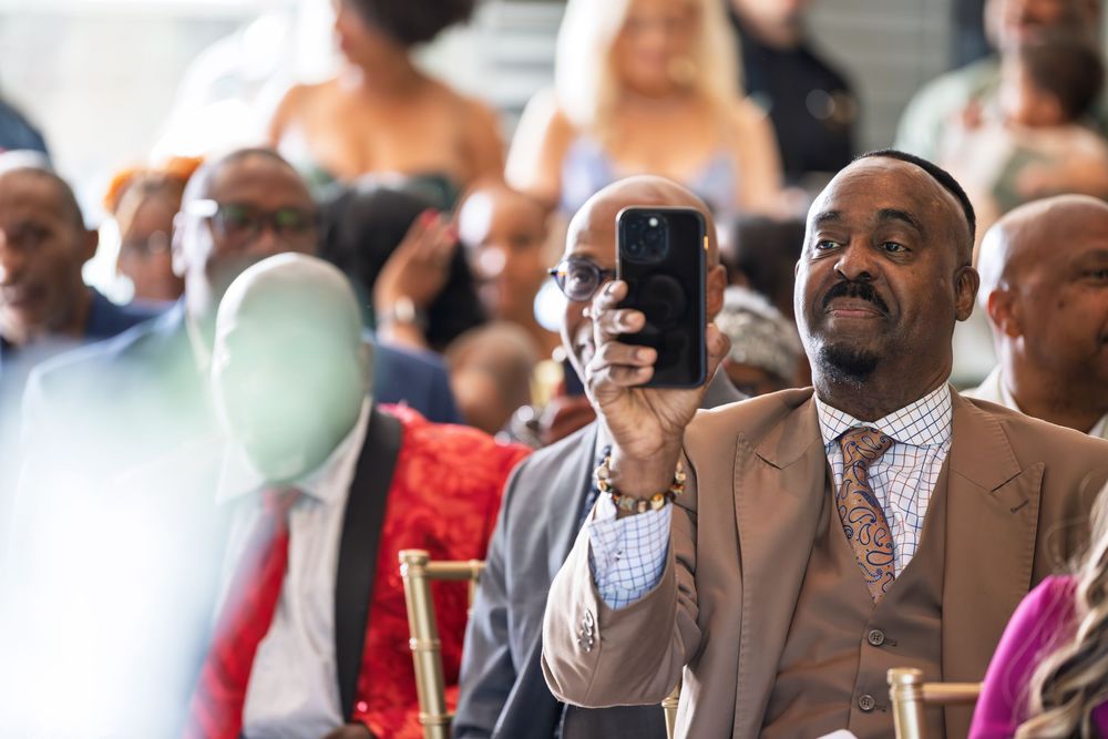 A man in a tan suit smiles while taking a photo with his smartphone at a formal event with guests seated behind him.