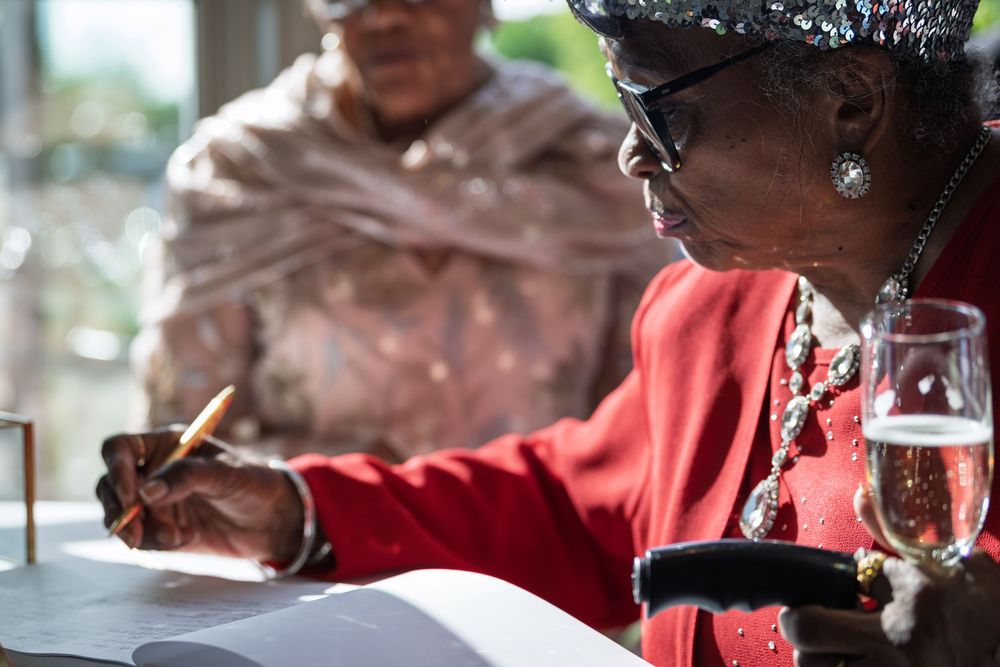 Elderly woman in red jacket signing documents at a table with champagne glass nearby.