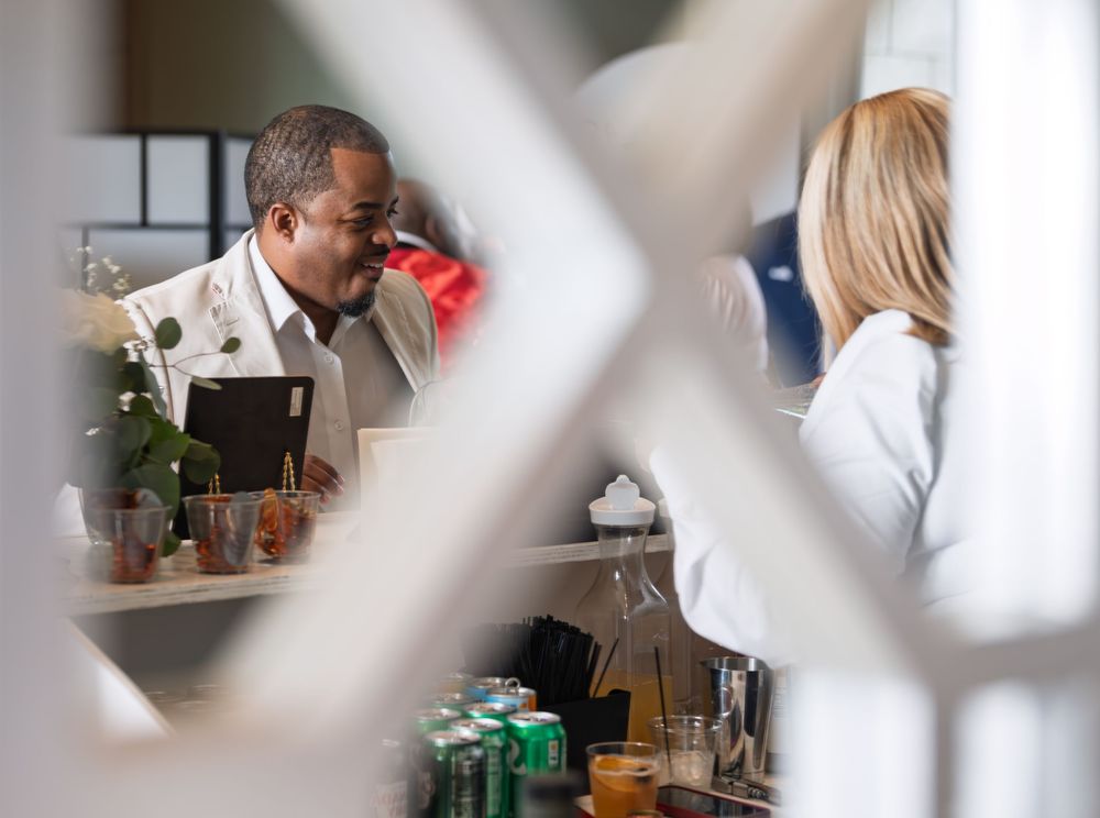 Two people in white coats converse at a bar counter with drinks and cans visible, viewed through a lattice window.