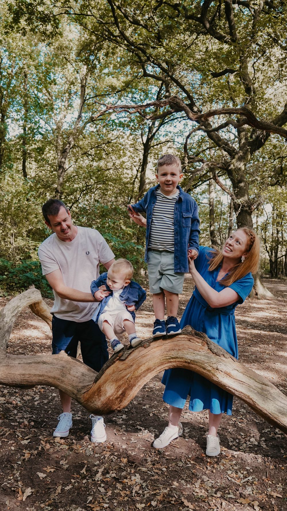 A family plays and laughs together while sitting on a fallen tree trunk in a wooded area.