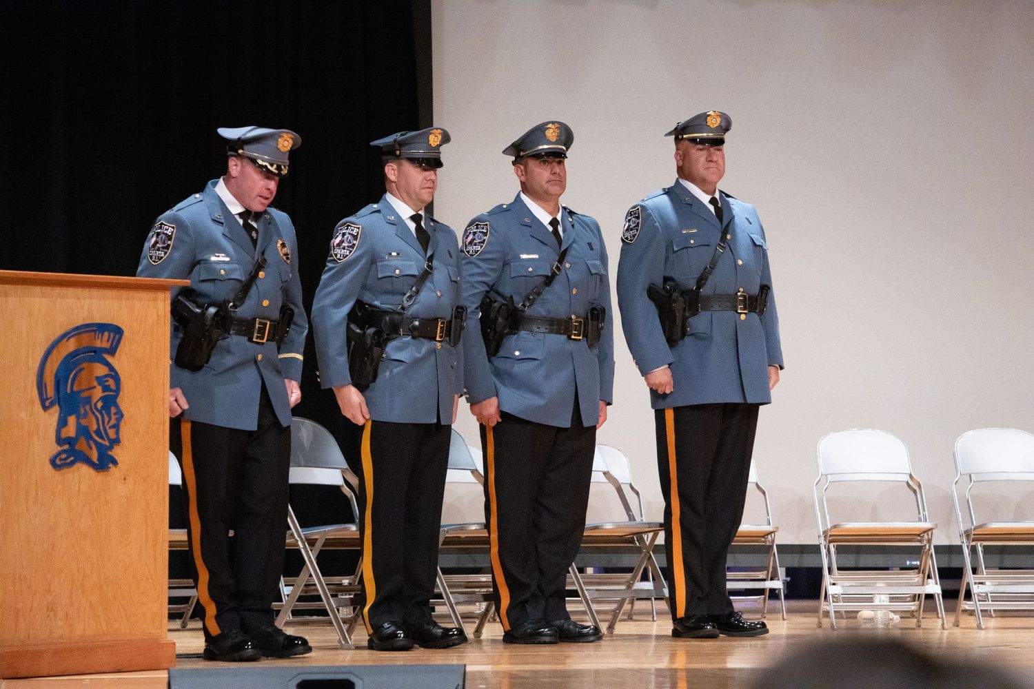 Four police officers in dress uniforms stand at attention on a stage during a formal ceremony.