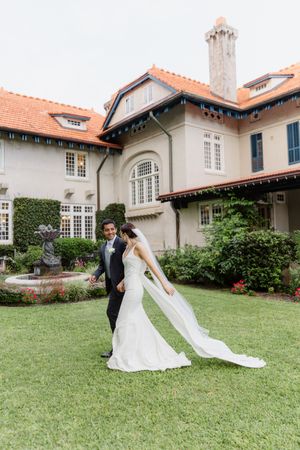 Bride in a white gown with a flowing veil walks hand in hand with her groom across a green lawn at a historic estate wedding.