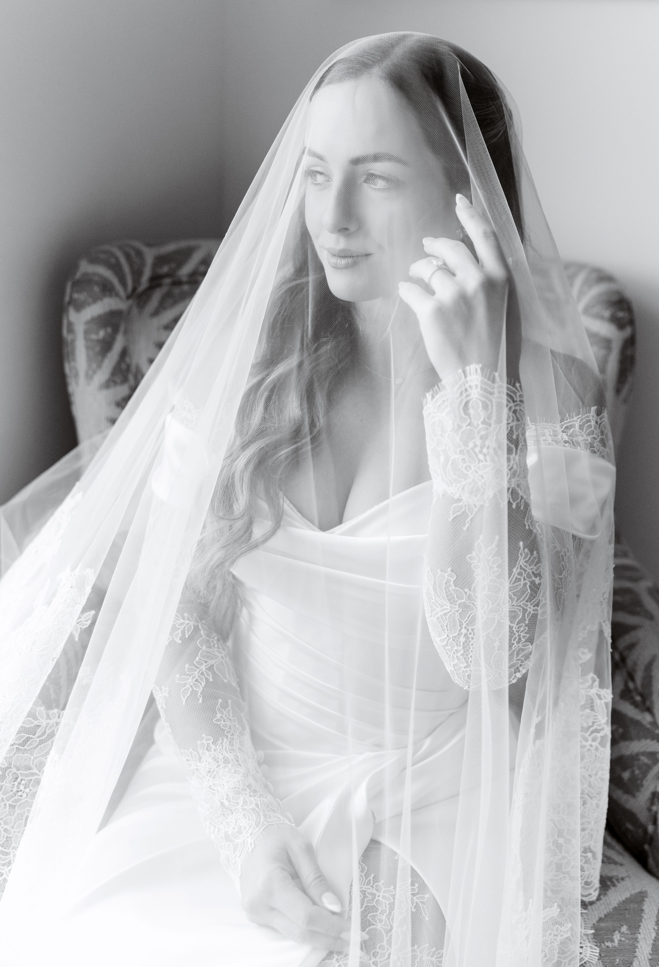Black and white bridal portrait of a bride draped in a lace-trimmed veil, seated gracefully in soft natural light.