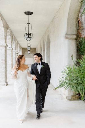 Bride and groom walk hand-in-hand through elegant arched corridor, she in white gown, he in black tuxedo.