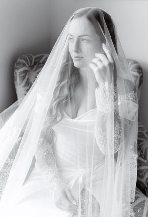 Black and white bridal portrait of a bride draped in a lace-trimmed veil, seated gracefully in soft natural light.