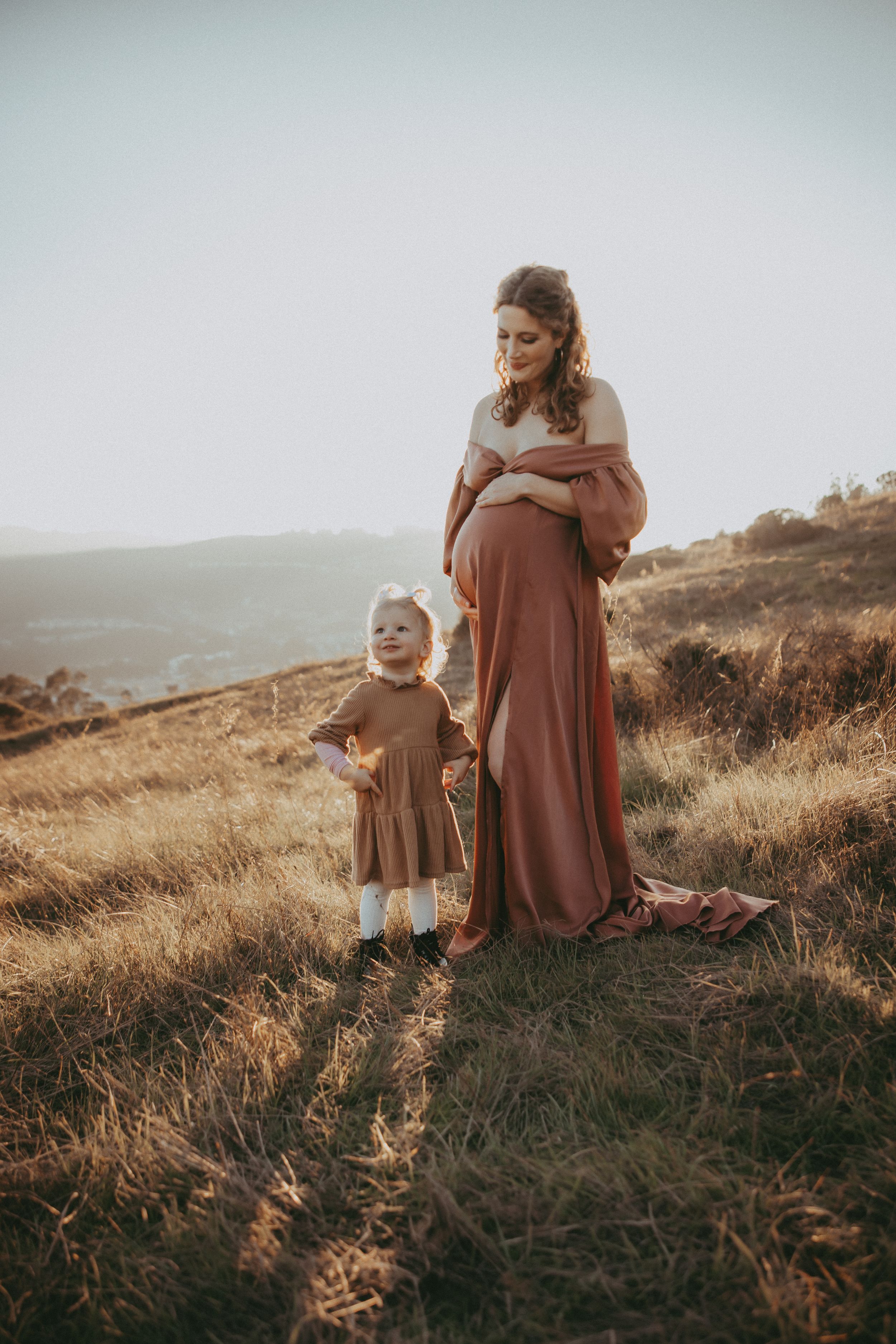 Pregnant woman in rust maxi dress with toddler girl on golden hillside at sunset.